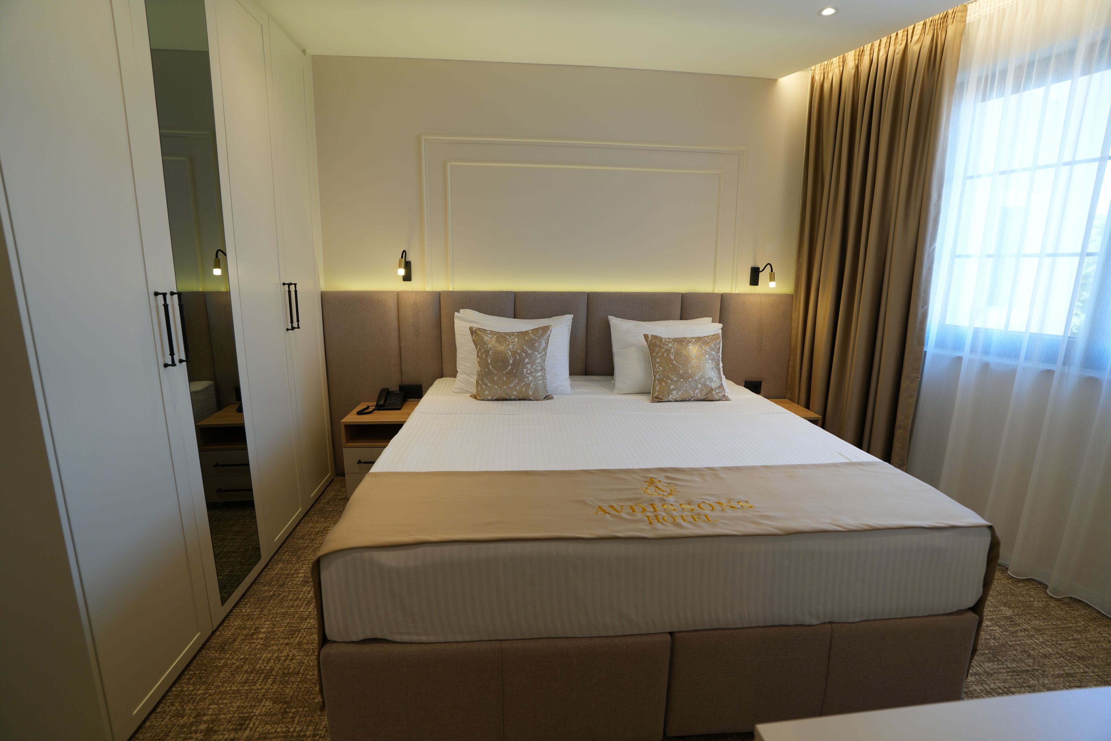 A hotel room with a large bed, a wardrobe, a bedside table, and a window with curtains.