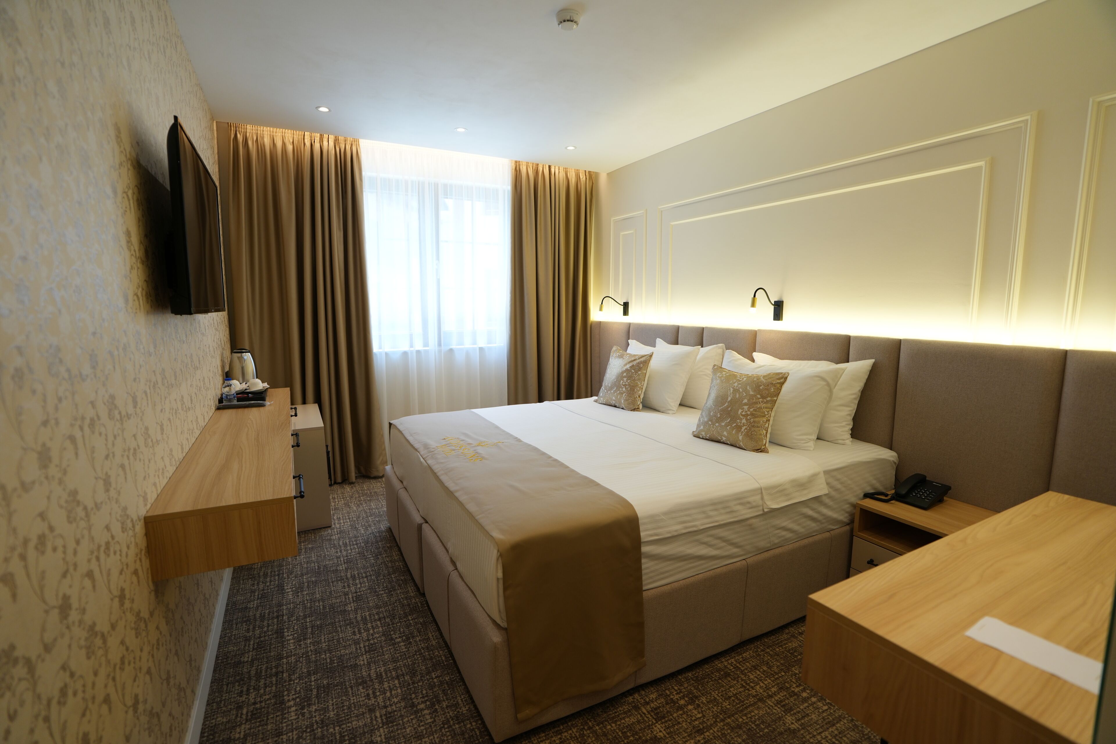 A hotel room with a large bed, bedside tables, a flat-screen TV, a desk, and a window with curtains.