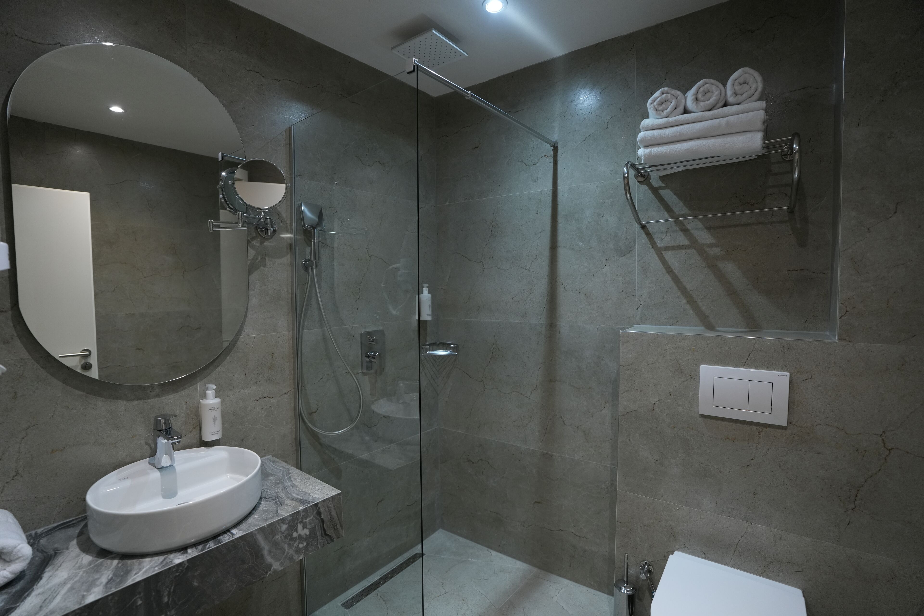 A modern bathroom with a glass-enclosed shower, a round mirror, a marble sink, and a towel rack with folded towels.