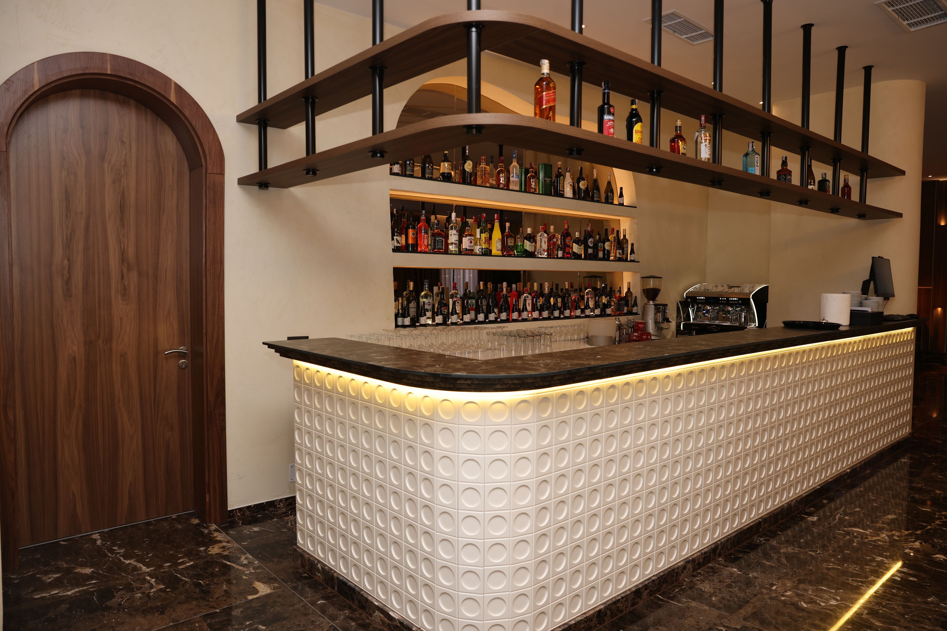 A bar area with a marble floor, a wooden door, and shelves with bottles.