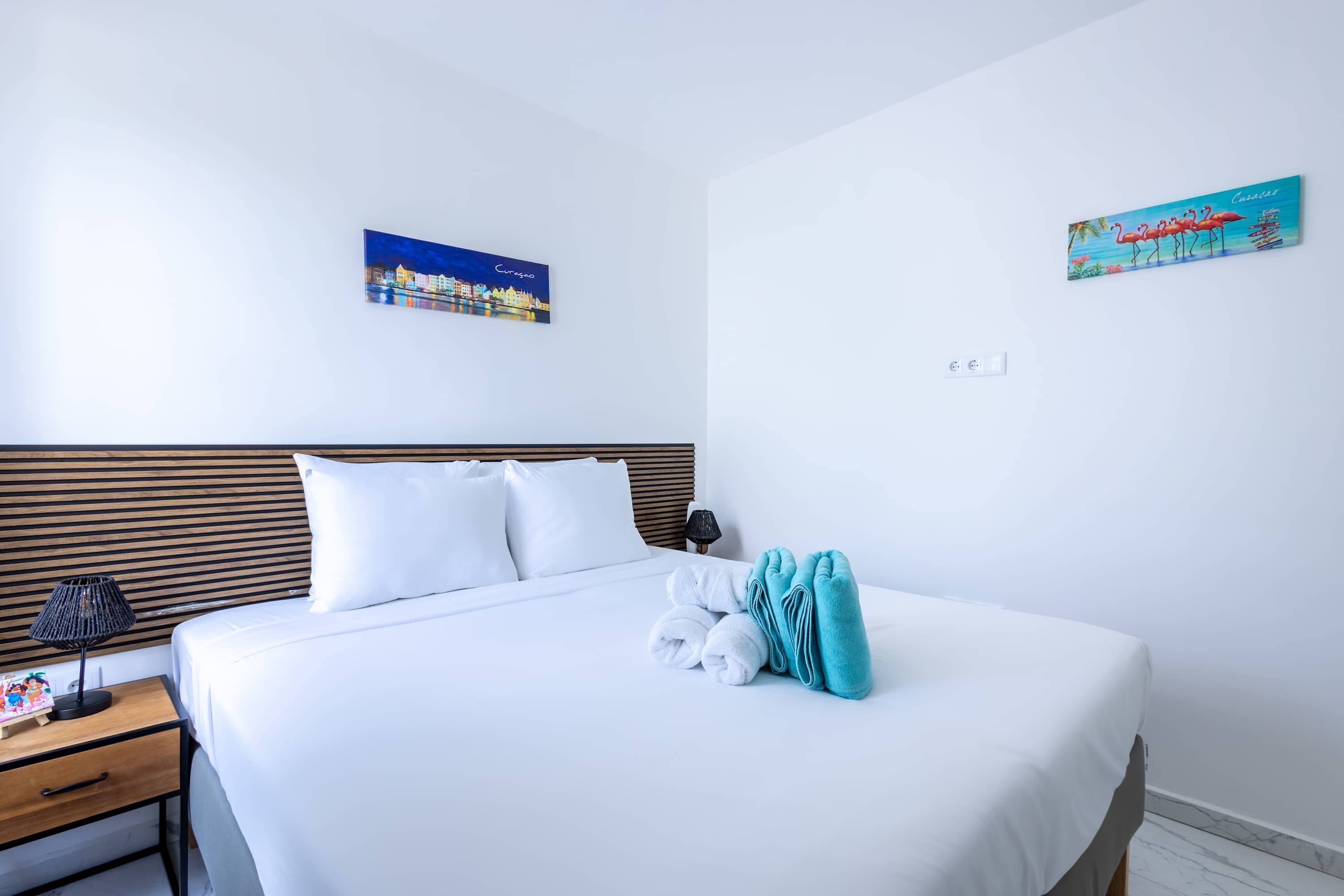 A hotel room with a neatly made bed, a nightstand with a lamp, and two wall art pieces depicting a cityscape and flamingos.