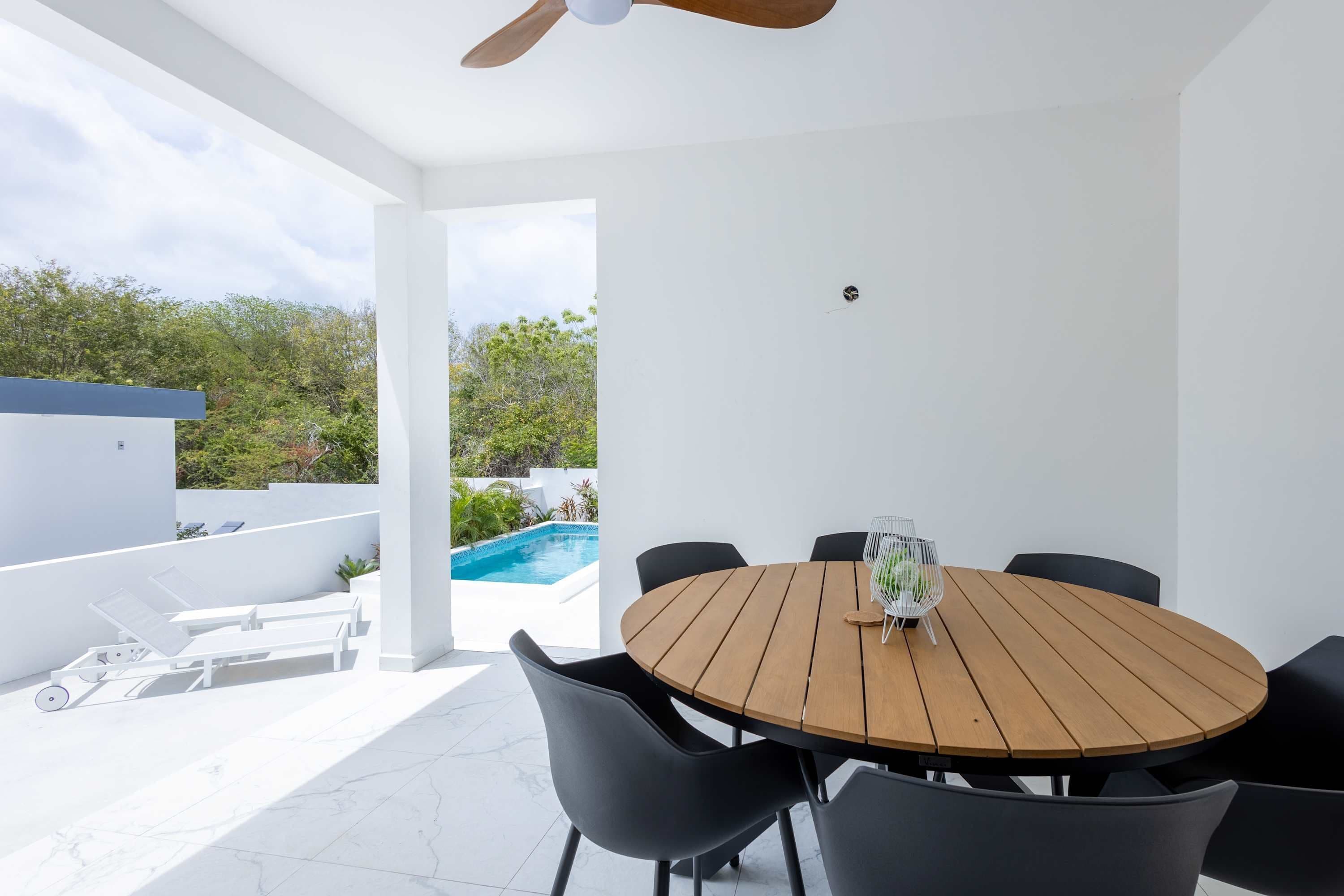 Un espacio moderno para comer al aire libre con mesa de madera y sillas negras, con vistas a una piscina y abundante vegetación.