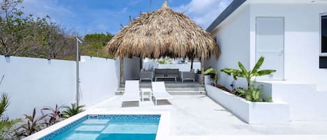 A modern outdoor pool area with a thatched roof structure, a white building, and a clear blue sky.