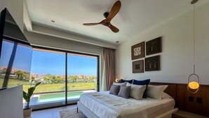 A modern bedroom with a large bed, a ceiling fan, and a view of a swimming pool and buildings. - Marina Garden 2 Bedroom Apartment With Picuzzi (Punta Cana)