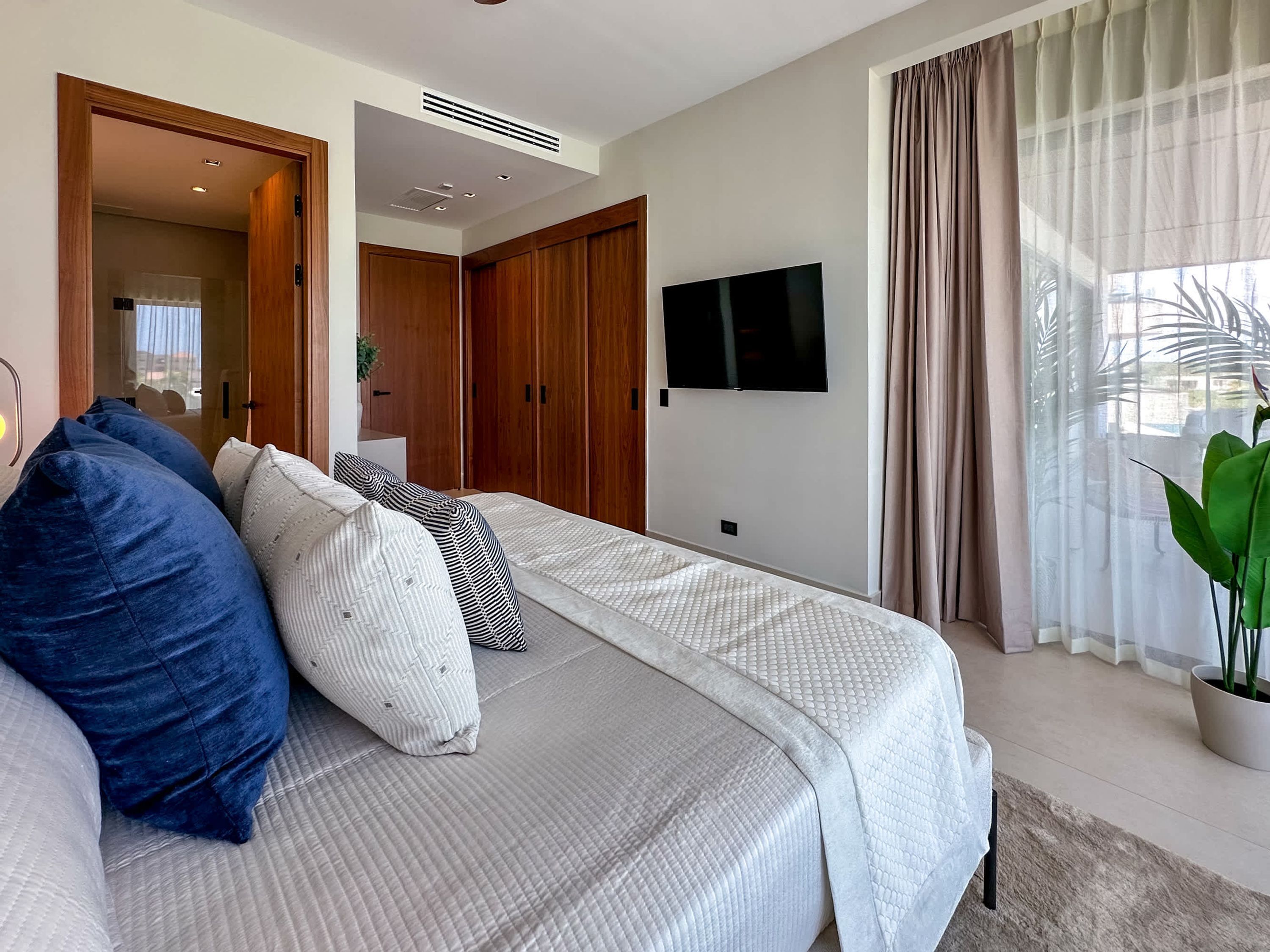 A modern bedroom with a large bed, a TV mounted on the wall, and a view of the outdoors through sheer curtains.