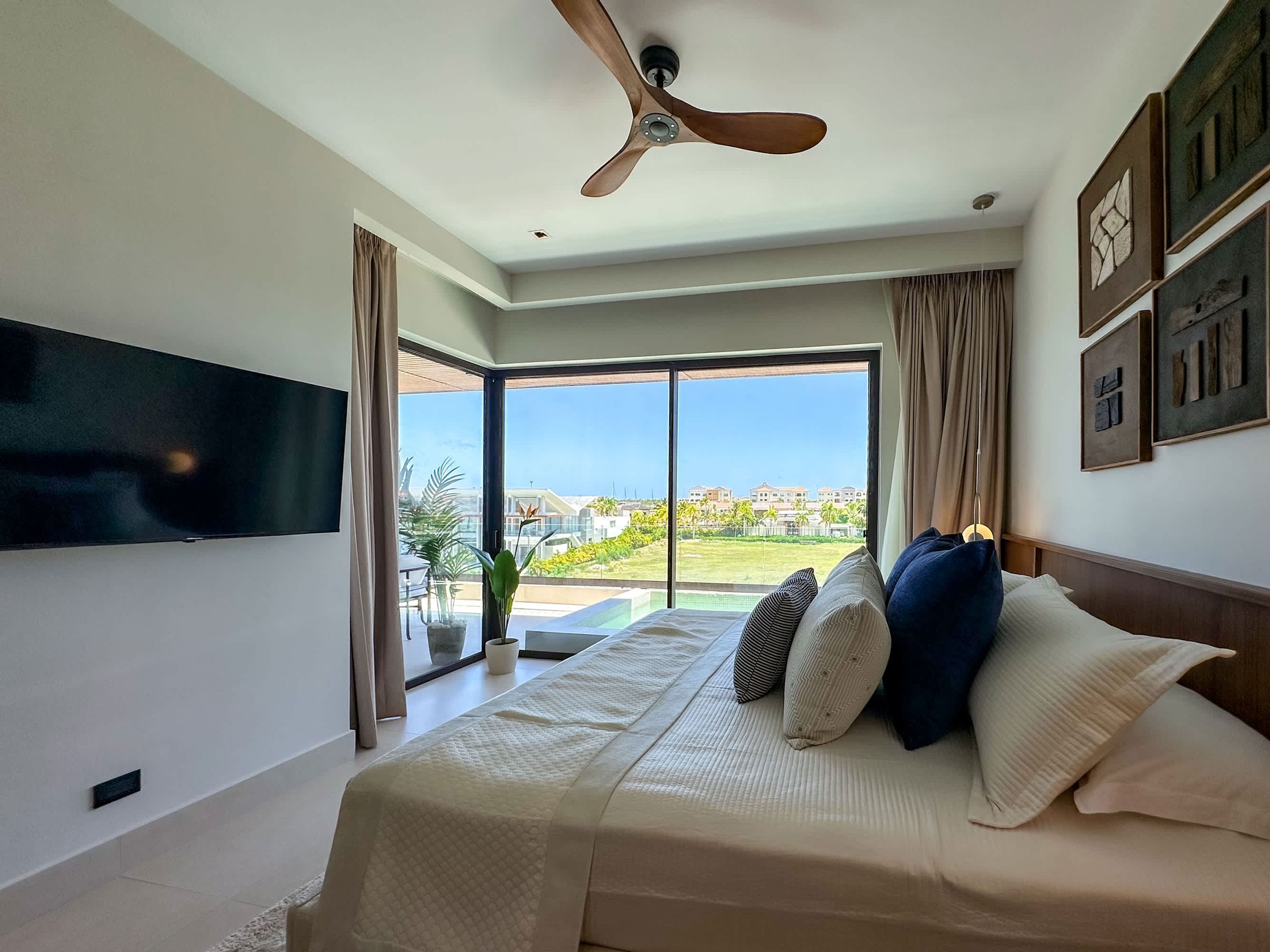 A bedroom with a large bed, a ceiling fan, a TV mounted on the wall, and a view of a residential area through large windows.