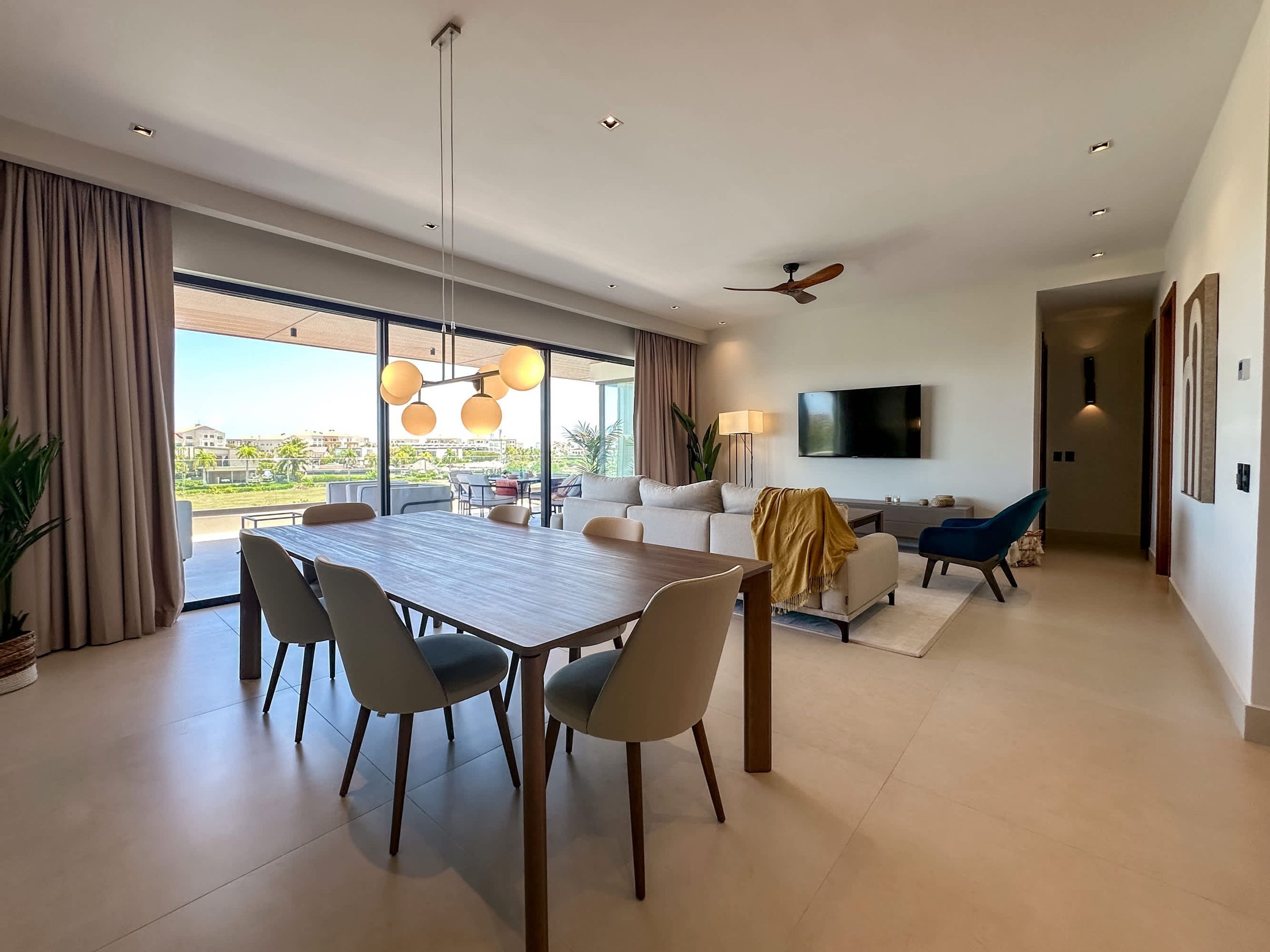 A modern living room with a dining area, large windows, a flat-screen TV, and a ceiling fan.