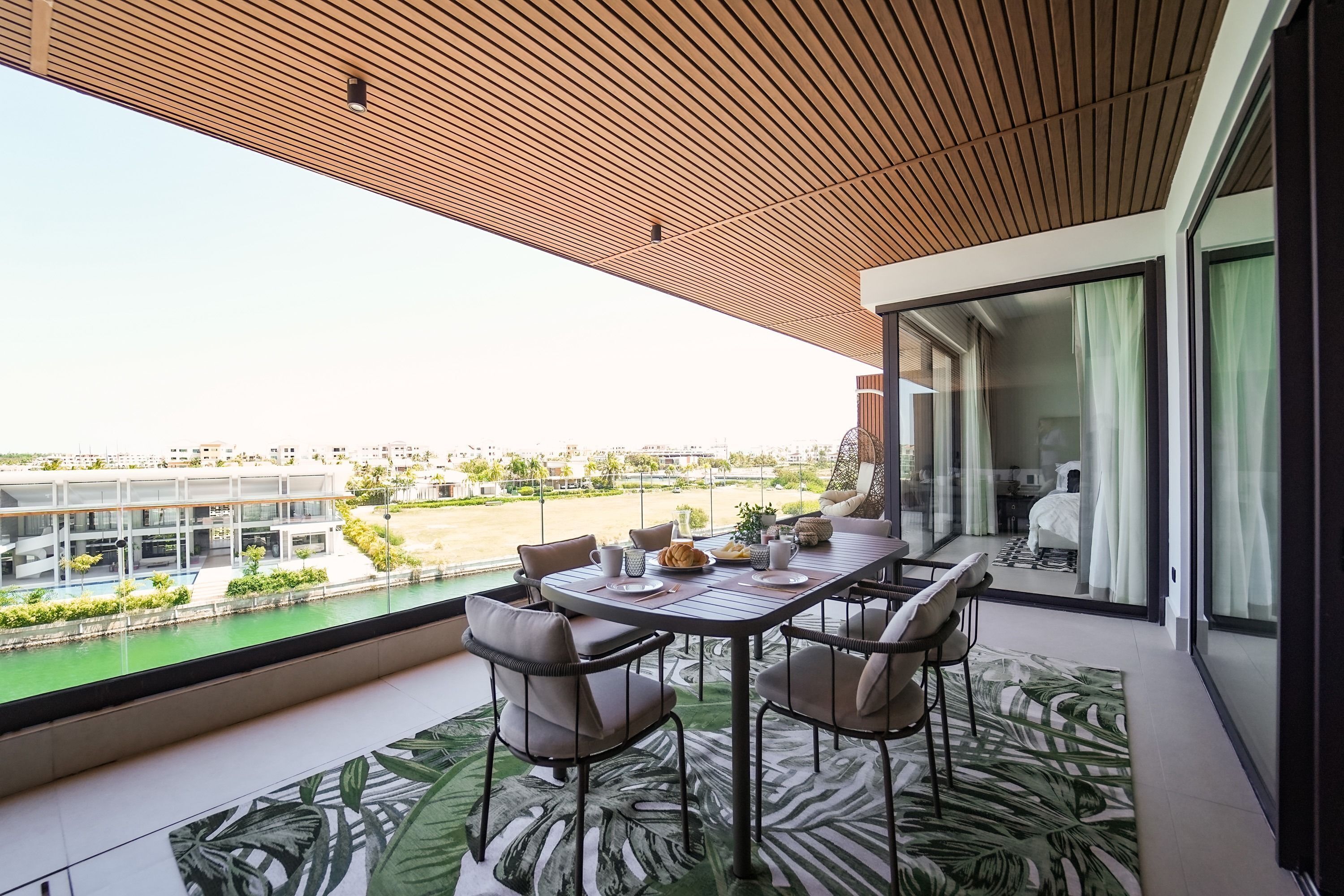 Ein Balkon mit Tisch und Stühlen, Blick auf einen Fluss und Gebäude, sowie ein modernes Interieur mit einem gemusterten Teppich.