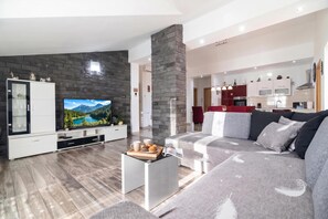 Un salon moderne avec un téléviseur à écran plat, un mur d’accent en pierre, un canapé gris et une cuisine en arrière-plan.