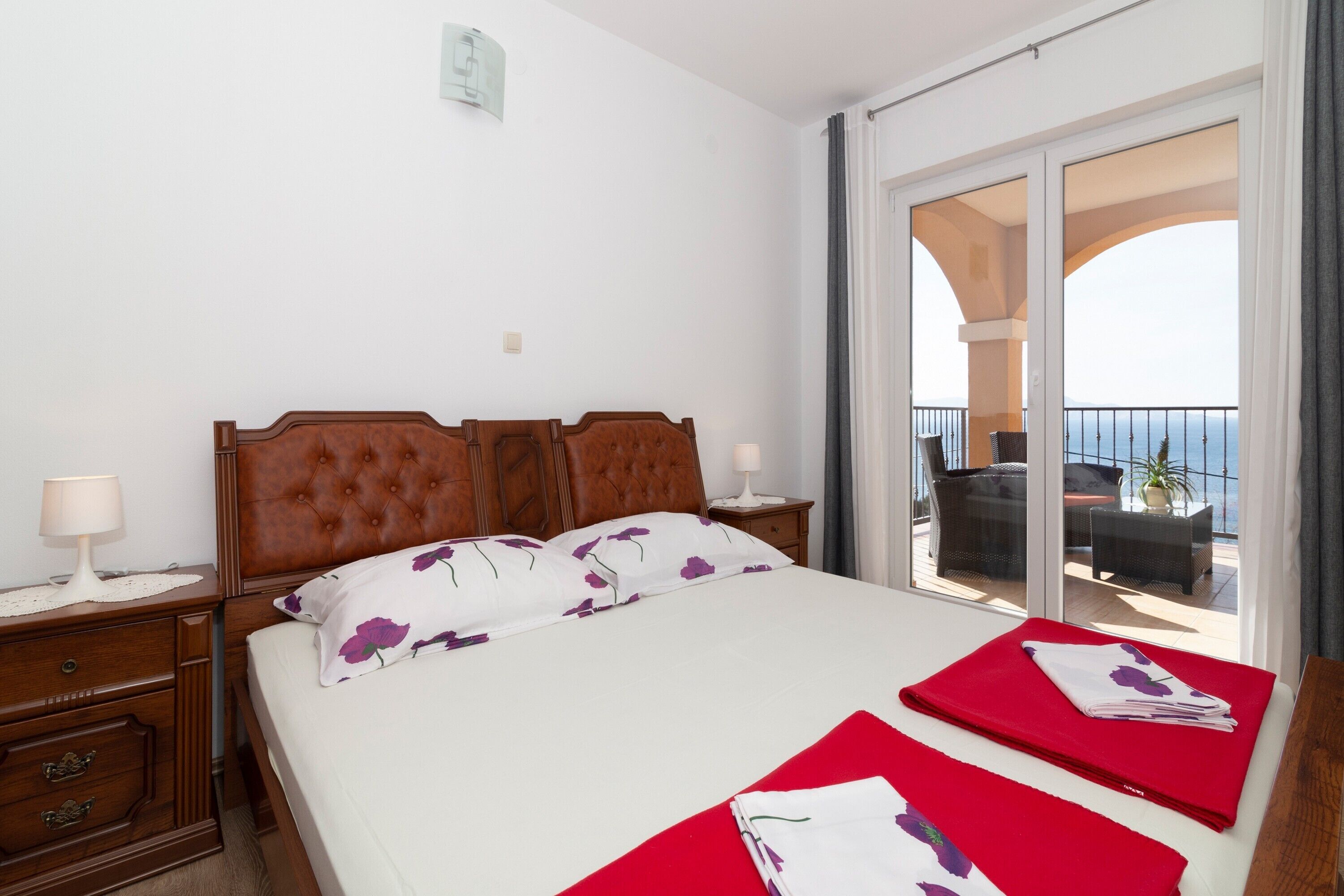 Hotelkamer met een groot bed, houten hoofdeinde, twee nachtkastjes met lampen, een balkon met uitzicht op zee, en een schuifdeur van glas.