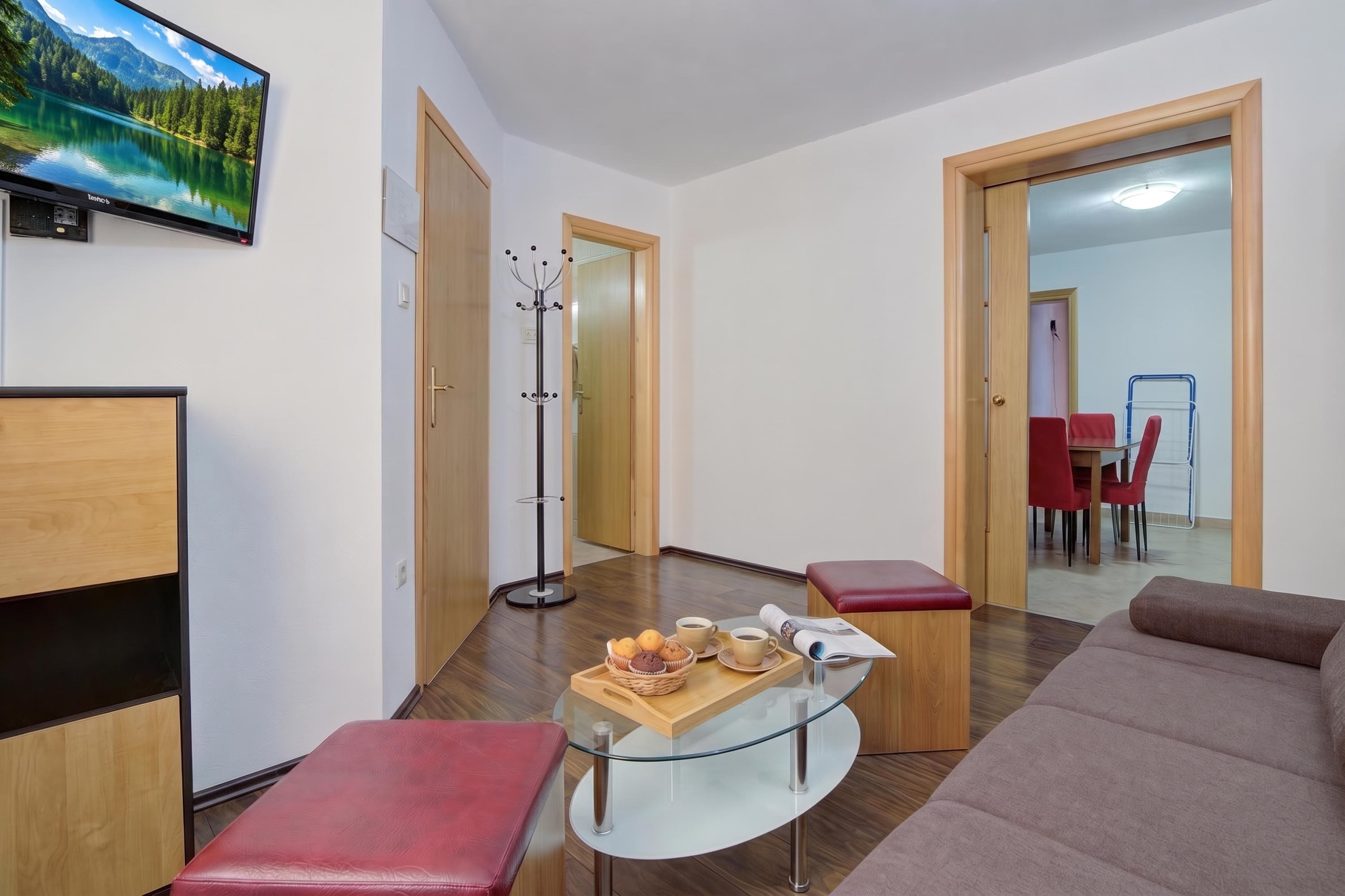 A modern living room with a glass coffee table, red leather bench, and a sofa. There is a coat rack and a television mounted on the wall.