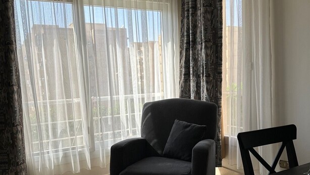A room with a large window, a black armchair, a dining table, and a patterned wall. - Spacious 3-Bd Hotel Apt in Gated Compound in Sheikh Zayed (Giza Governorate)
