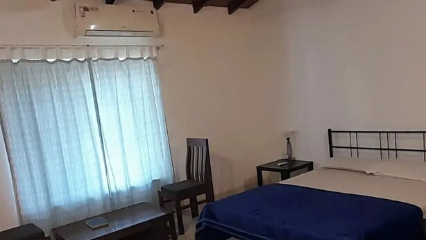 Ein Schlafzimmer mit einem Bett, einem Sessel, einem kleinen Tisch, einer Klimaanlage und einem Fenster mit Vorhängen.