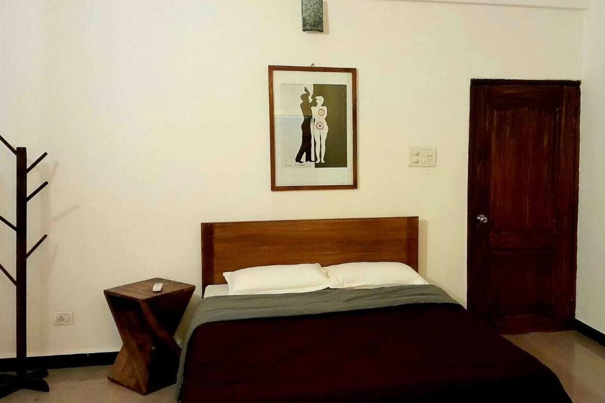 Un dormitorio con cabecera de madera, cama con ropa de cama blanca y gris, un cuadro enmarcado en la pared, una puerta de madera y una mesita de noche de madera con una lámpara.