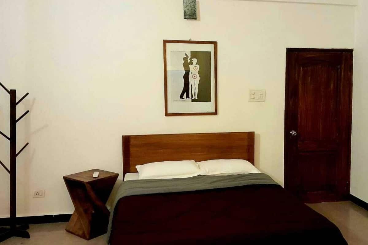 A bedroom with a wooden headboard, a bed with white and gray bedding, a framed picture on the wall, a wooden door, and a small wooden nightstand with a lamp.