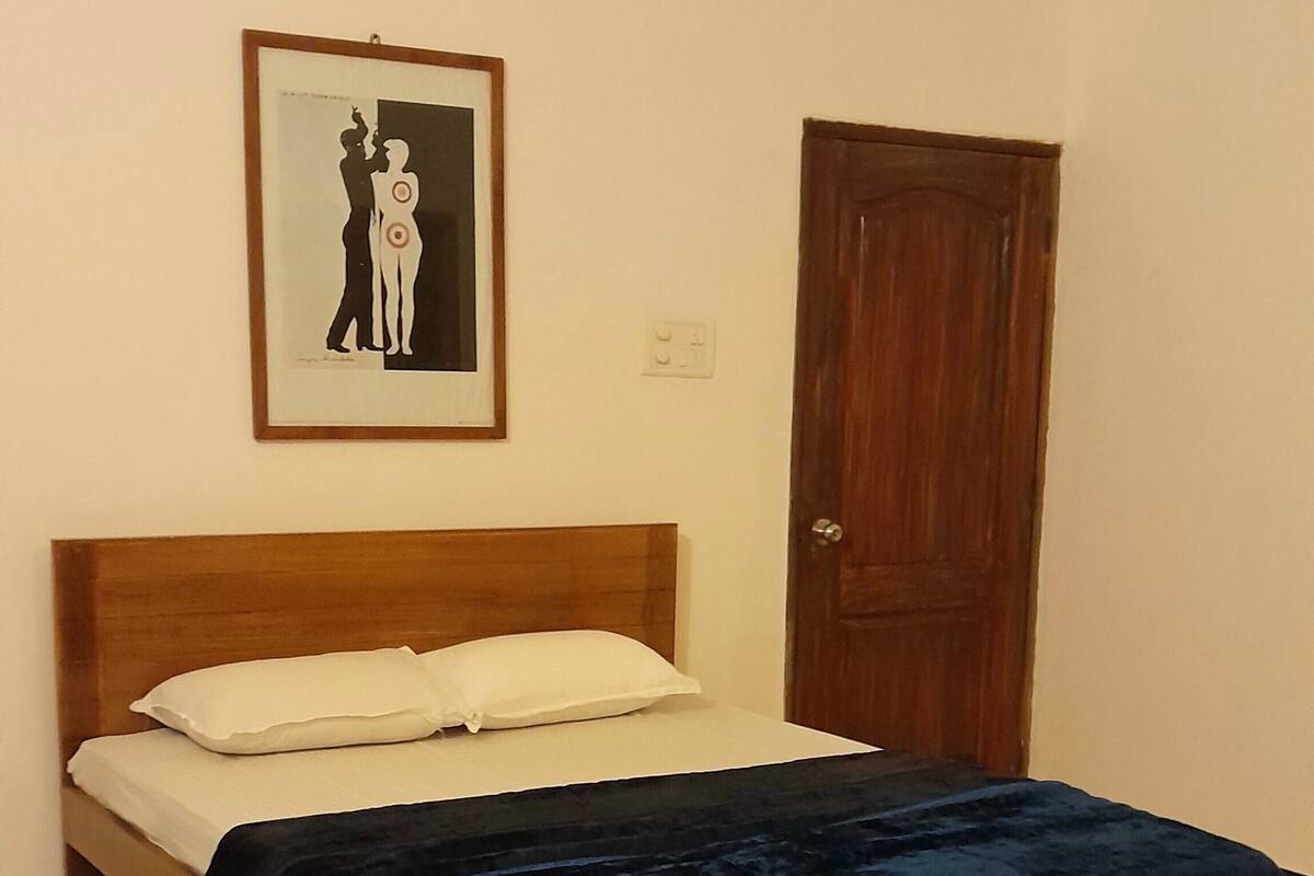 Una cama individual con cabecera de madera, almohada blanca y manta azul. Encima de la cama, un cuadro enmarcado cuelga de la pared.