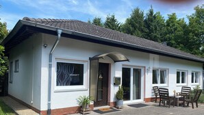 Een laag gebouw met pannendak, een voordeur en een klein voortuintje met twee stoelen en een tafel.