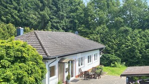 Een wit gebouw met een pannendak, een terras met tuinmeubilair en een weelderig groen bos op de achtergrond.
