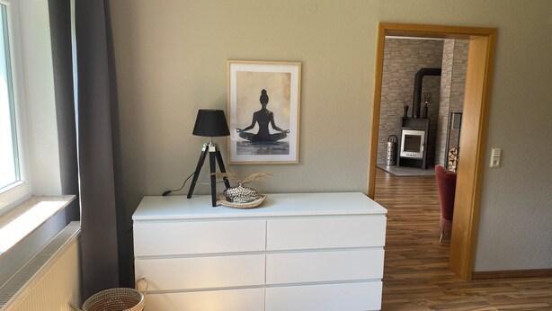 Een kamer met een witte dressoir, een lamp op een statief, een lijst met een foto aan de muur, een mand en een deur naar een andere kamer.