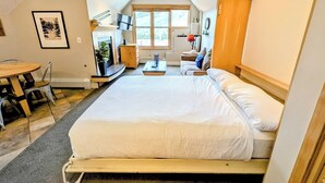 A bedroom with a large bed, a desk, and a chair. There is a window with a view of trees.
