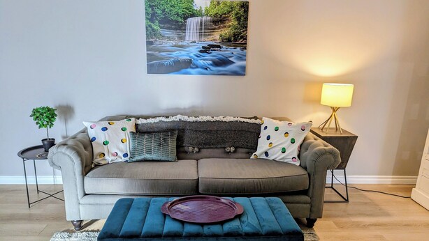 Un salon avec un canapé gris, un pouf bleu, une petite table d’appoint avec une lampe, et un tableau encadr é représentant une cascade accroché au mur.