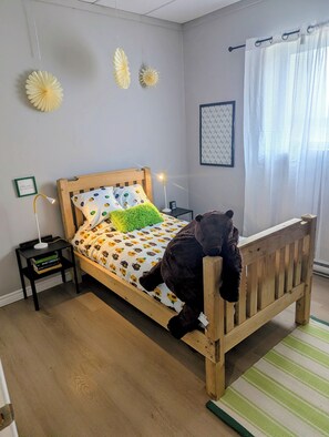 A bedroom with a wooden bed, a green pillow, a teddy bear, a nightstand with a lamp, and a framed picture on the wall.
