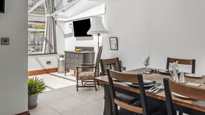 A dining area with a wooden table set for two, a chair with a cushion, a potted plant, a window with curtains, a cabinet, and a wall-mounted lamp. - Stylish Barn Getaway in Carmarthenshire (Carmarthen)