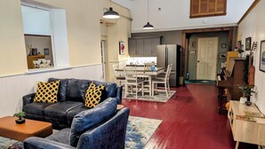 A living room with a dark blue sofa, a coffee table, and a patterned rug. There is a dining area with a white table and chairs, and a kitchen with a refrigerator and cabinets.