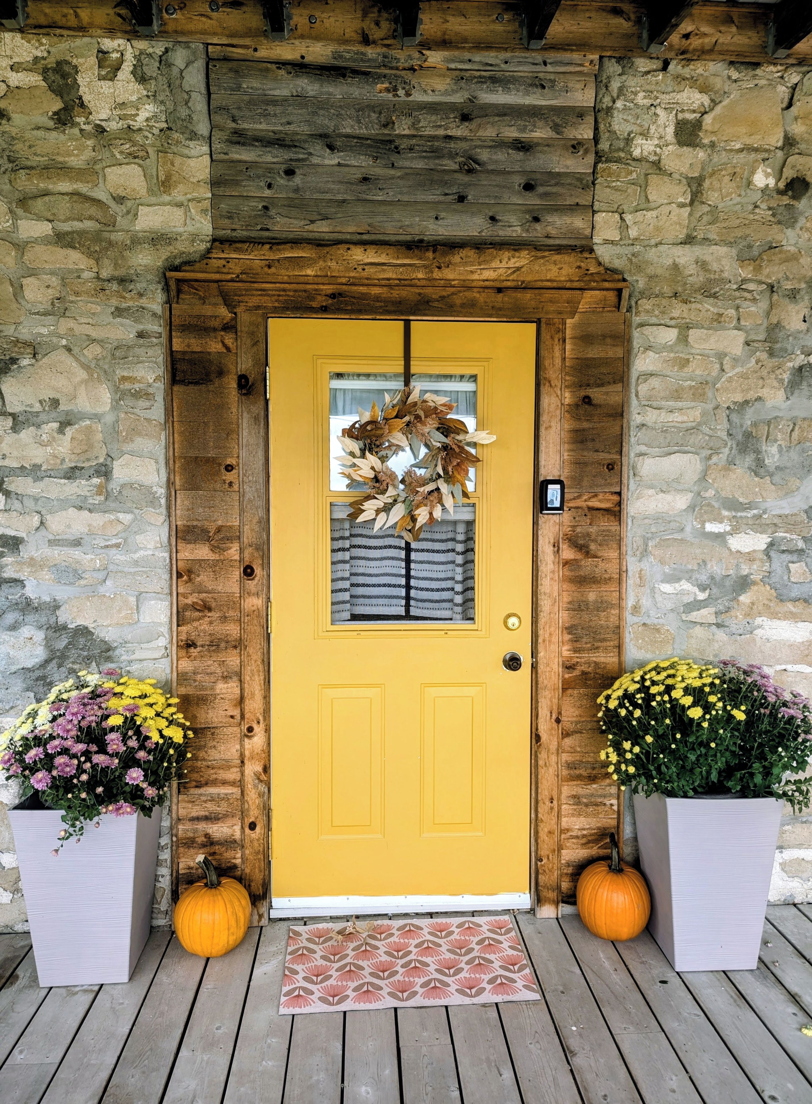 A yellow door with a wreath, flanked by two potted plants with flowers, and a wooden porch with a patterned mat.