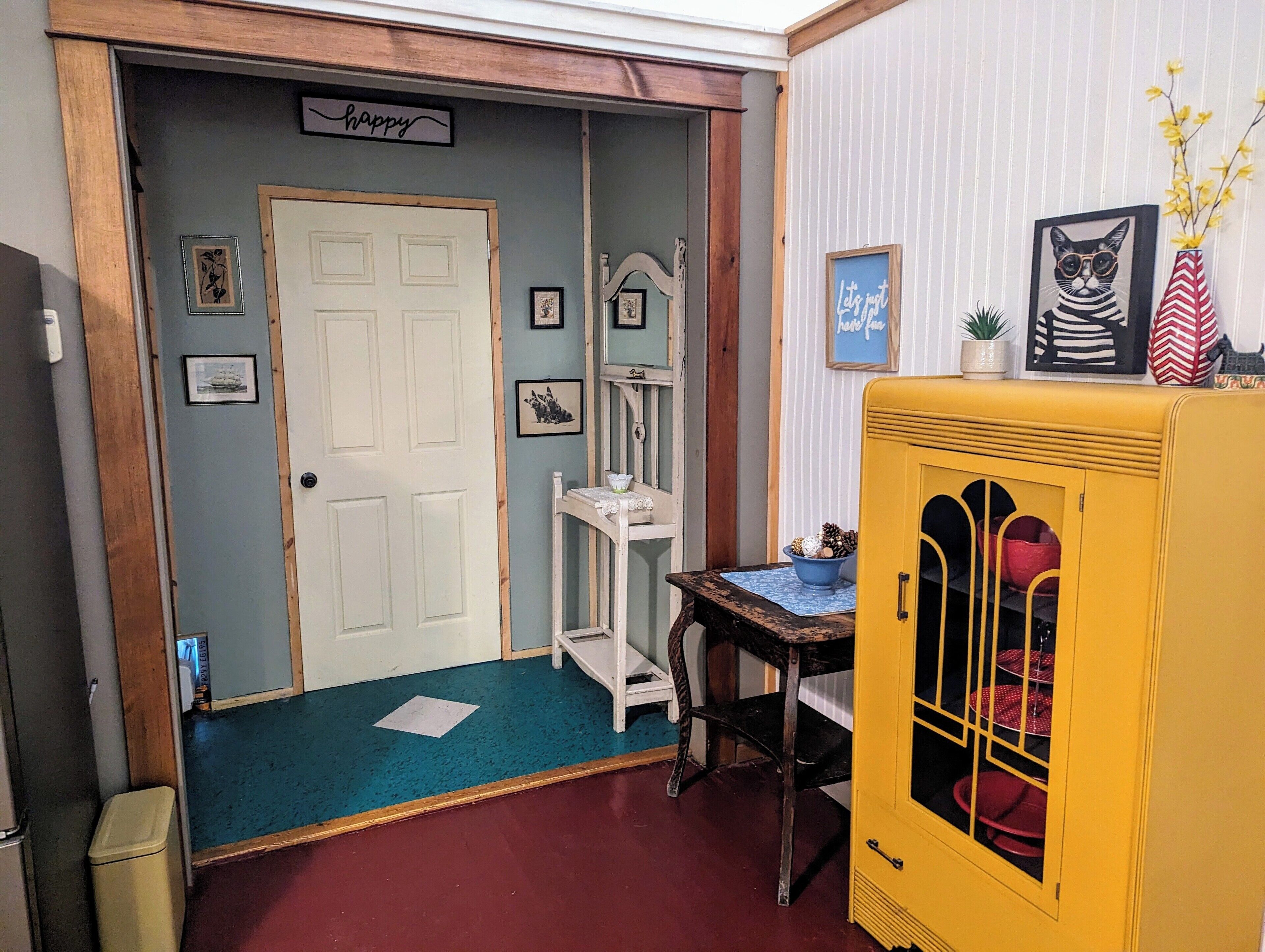 A room with a white door, a small table with a blue tablecloth, a yellow cabinet with glass doors, and a mirror on the wall.