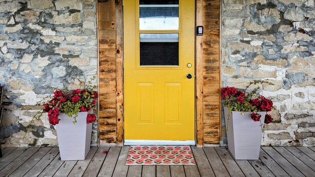 A yellow door with a window, flanked by stone walls and wooden beams, with two flower planters on either side.