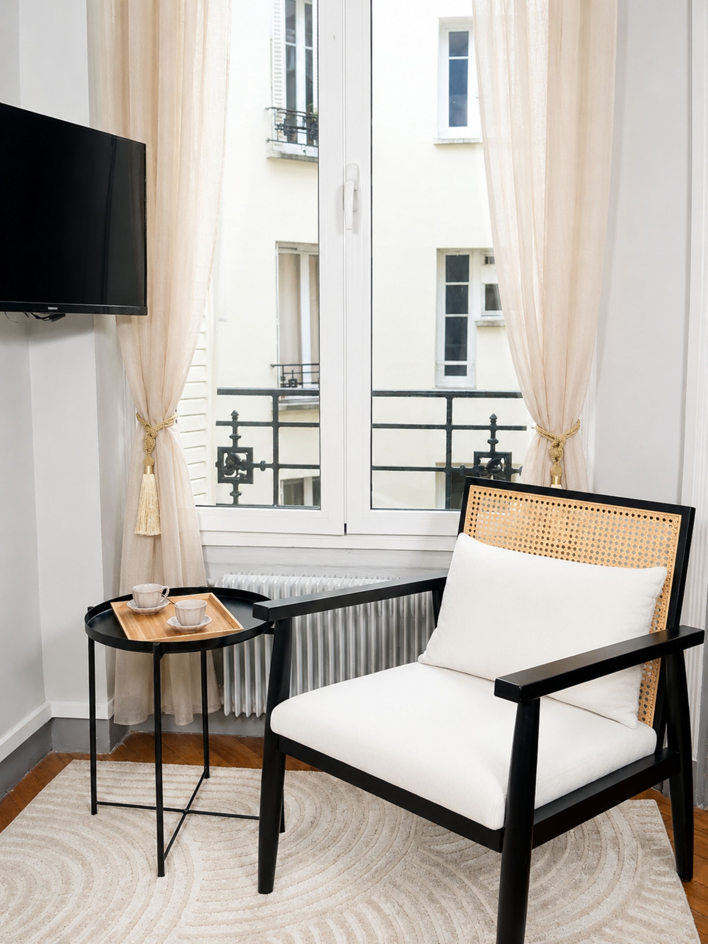 A modern room with a black chair, a small table with a tray, a TV mounted on the wall, and a window with curtains.