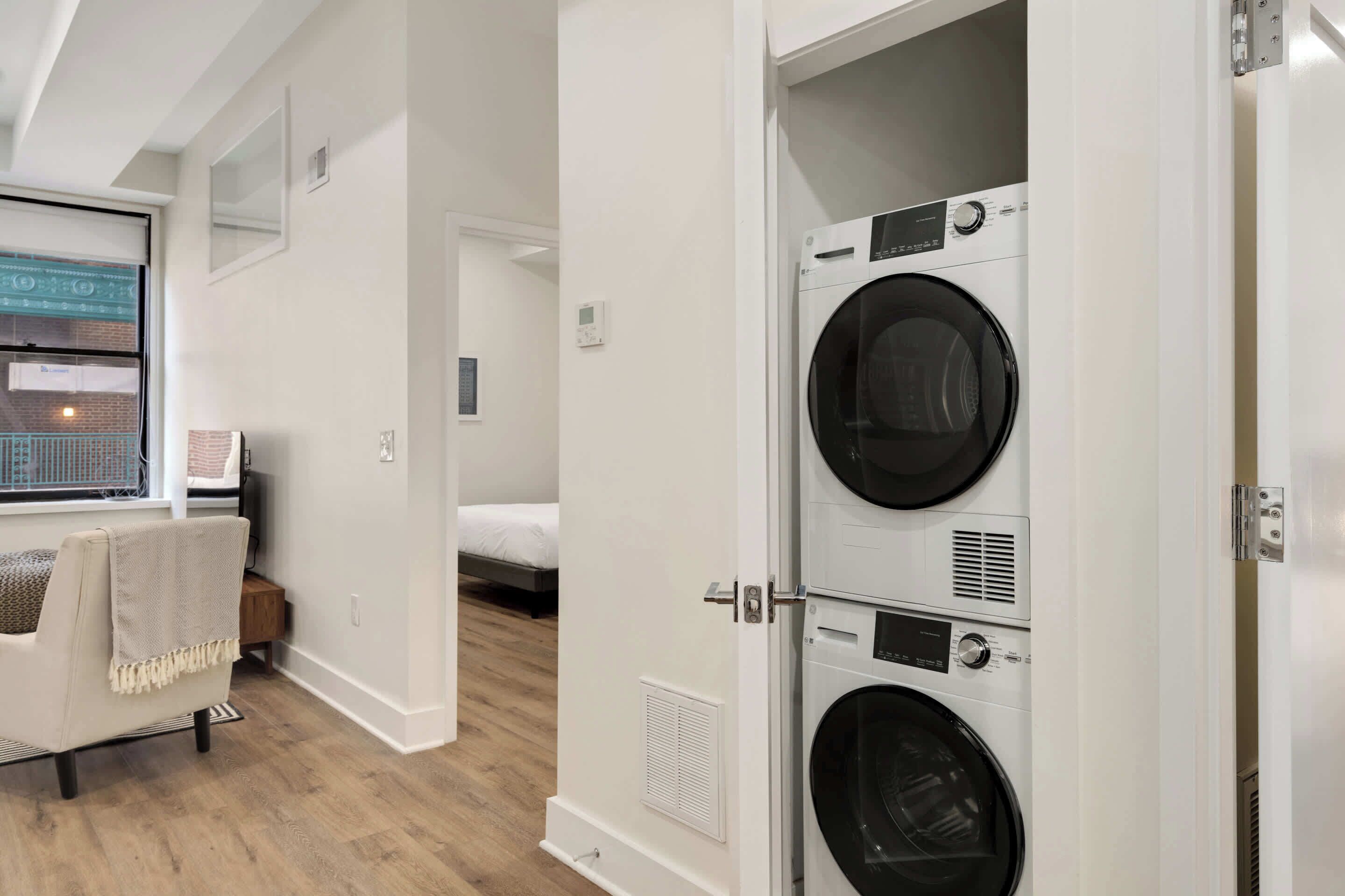 A laundry room with two stacked washing machines, a bed in the adjacent room, and a window with a view of a brick building.