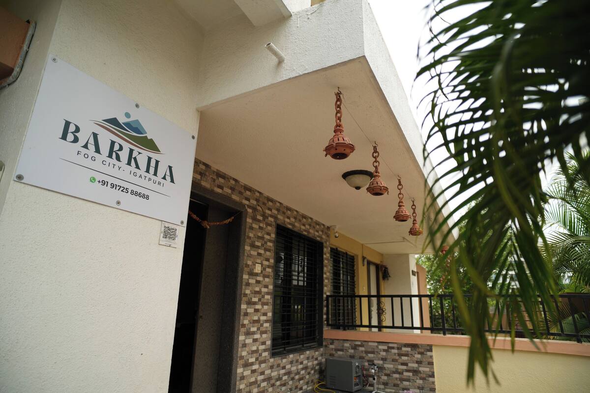 A building with a sign reading "Barkha Fog City - Igatpuri" and a contact number. There are hanging lights and a window with a metal grill.