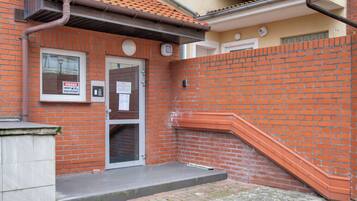 Een gebouw van baksteen met een dak van rode tegels, een witte deur en een klein buitenruimte met een metalen bank.