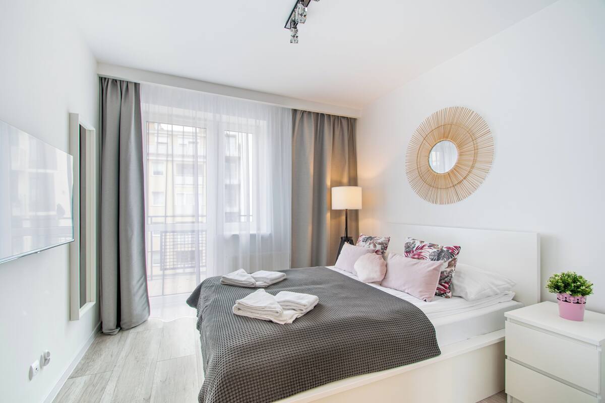 A modern hotel room with a large bed, a bedside table with a lamp, a wall-mounted mirror, and a window with curtains.