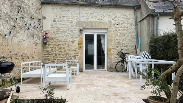 A stone building with a patio area featuring white furniture, a barbecue grill, and a bicycle. - The little sinner's house. (Arromanches-les-Bains)