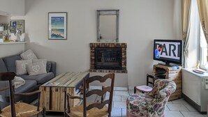 A living room with a fireplace, a television, a sofa, a coffee table, and a floral armchair. - The little sinner's house. (Arromanches-les-Bains)