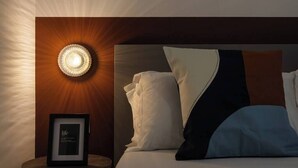 Un letto rifatto con lenzuola bianche e blu, un quadro incorniciato su un comodino e una lampada fissata al muro.