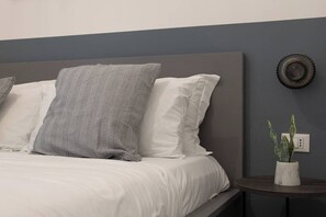 Un lit bien fait, avec des oreillers rayés blancs et gris, une tête de lit grise et une petite table ronde placée à côté du lit, sur laquelle se trouve une plante.