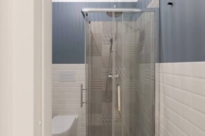 Une salle de bain moderne avec une douche à parois de verre, des murs recouverts de carreaux blancs et des toilettes apparentes.