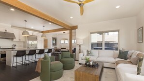 A modern kitchen with white cabinets, a bar area, and a dining space. There is a living area with a sofa, a coffee table, and a window with blinds.