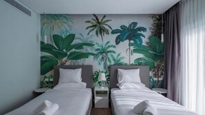 A hotel room with two single beds, a mural of tropical plants on the wall, a small table with a lamp, and a window with curtains. - Kennah Rivera | 2BR Garden Apt, Near EDNC Mall (New Cairo)