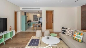 A modern living room with a sofa, a coffee table, and a TV on a stand. There is a kitchen in the background with a dining table and chairs. - Kennah Rivera | 2BR Garden Apt, Near EDNC Mall (New Cairo)
