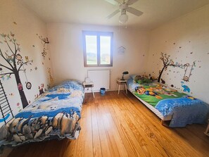 Quarto com duas camas de solteiro, janela, radiador, ventoinha no teto e uma pequena mesa com luminária.