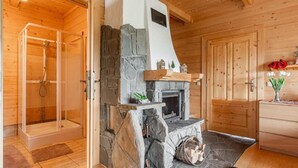 Interior de una cabaña de madera con chimenea de piedra, ducha con cerramiento de vidrio y puerta de madera.