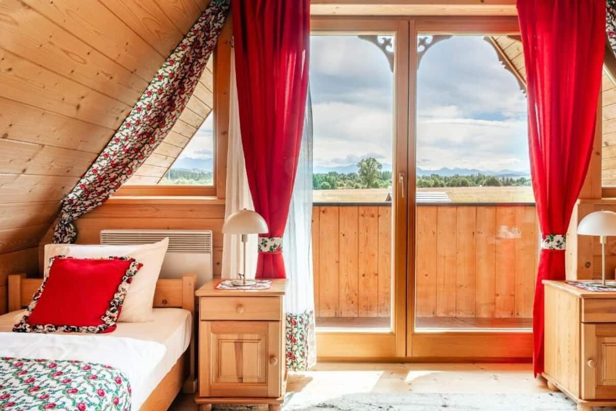 Un dormitorio acogedor con techo inclinado de madera, cama con ropa de cama roja y floral, y vista a un paisaje verde a través de un gran ventanal corredero.