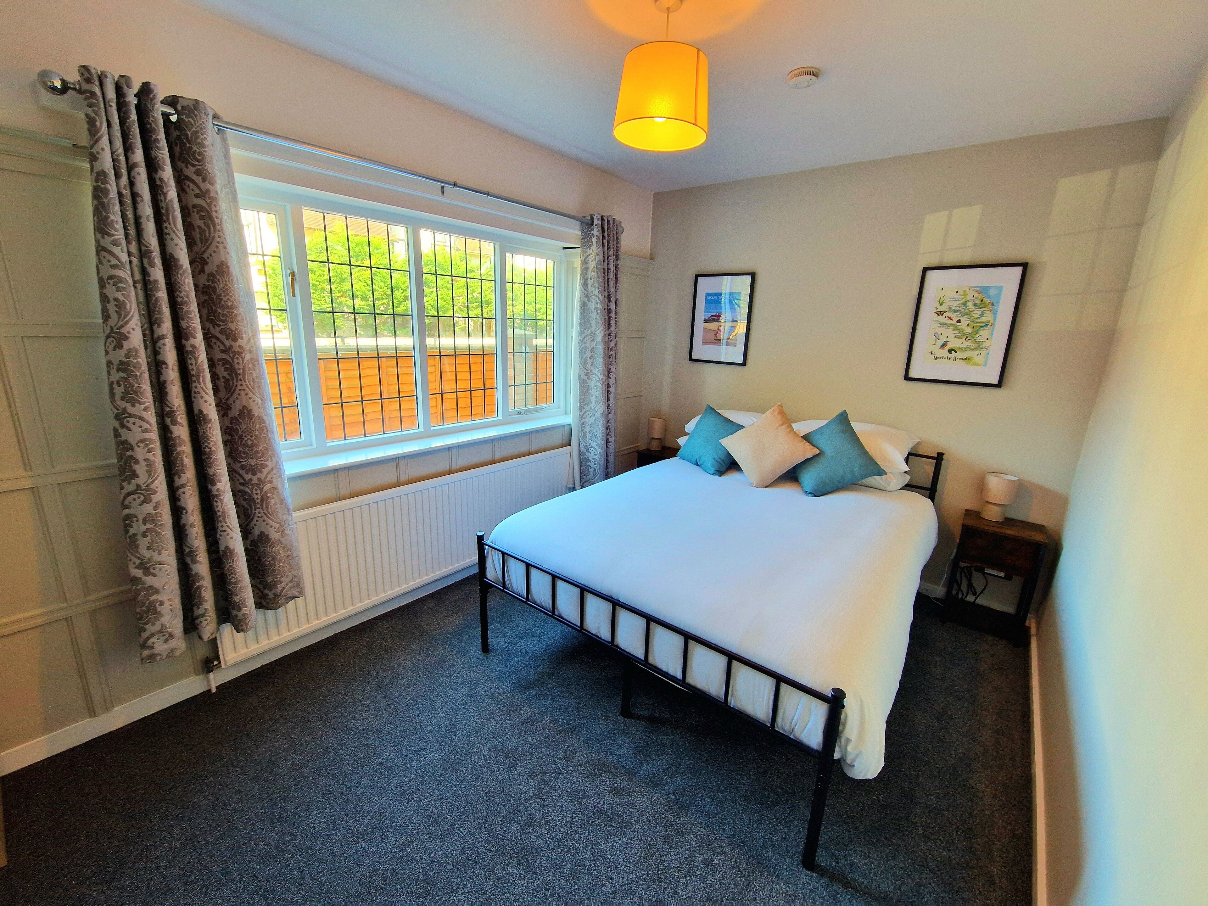 A bedroom with a bed, a window with curtains, a radiator, a bedside table with a lamp, and two framed pictures on the wall.