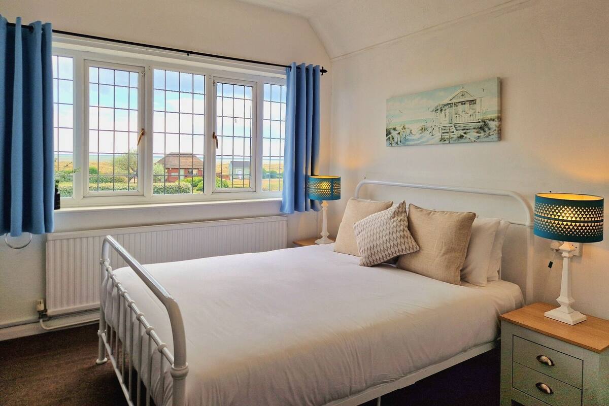 Une chambre à coucher avec un lit, deux tables de chevet avec des lampes, une fenêtre avec des rideaux, un radiateur et un tableau au mur.