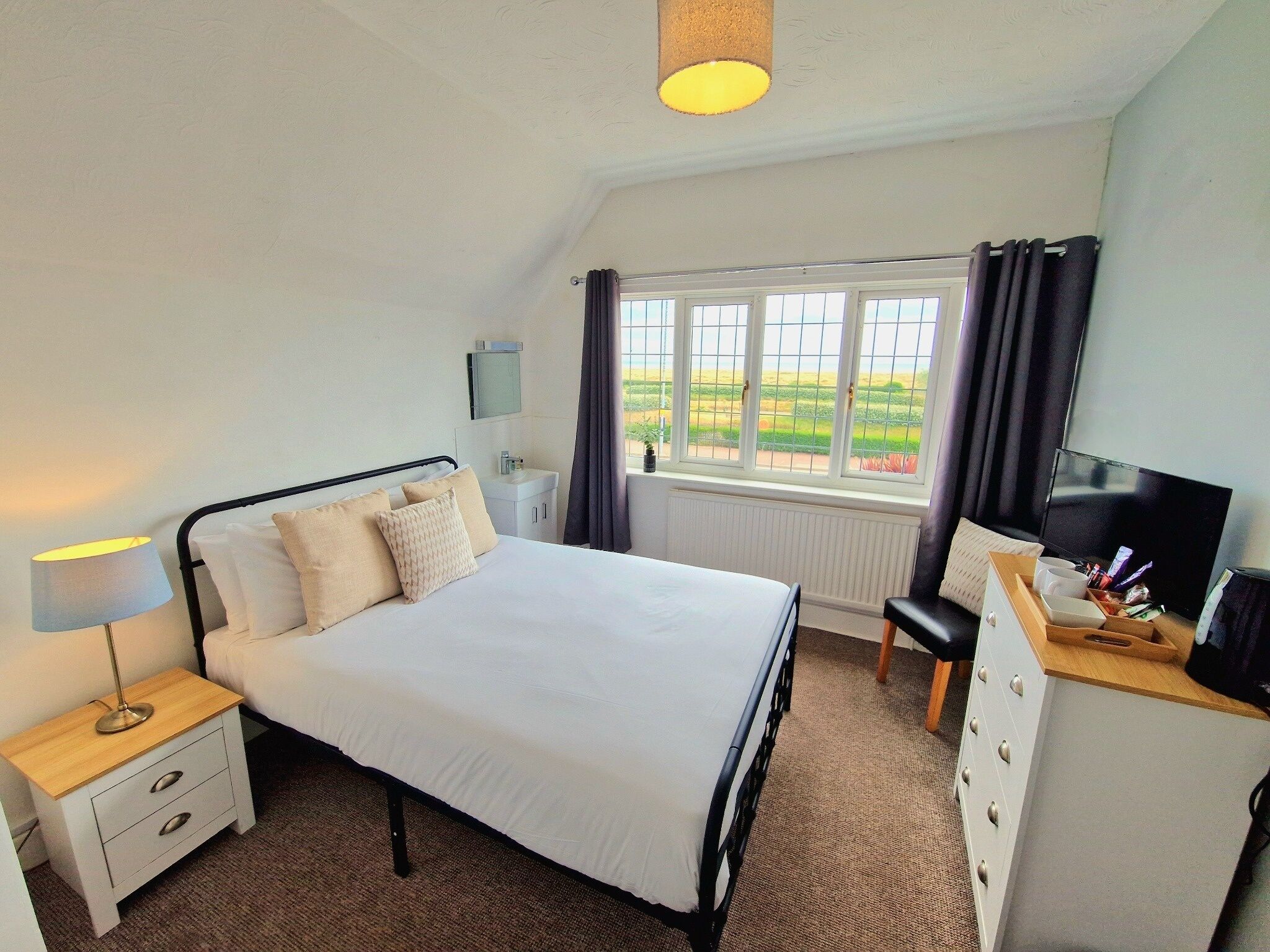 A bedroom with a bed, bedside table, lamp, TV, dresser, and a window with curtains.